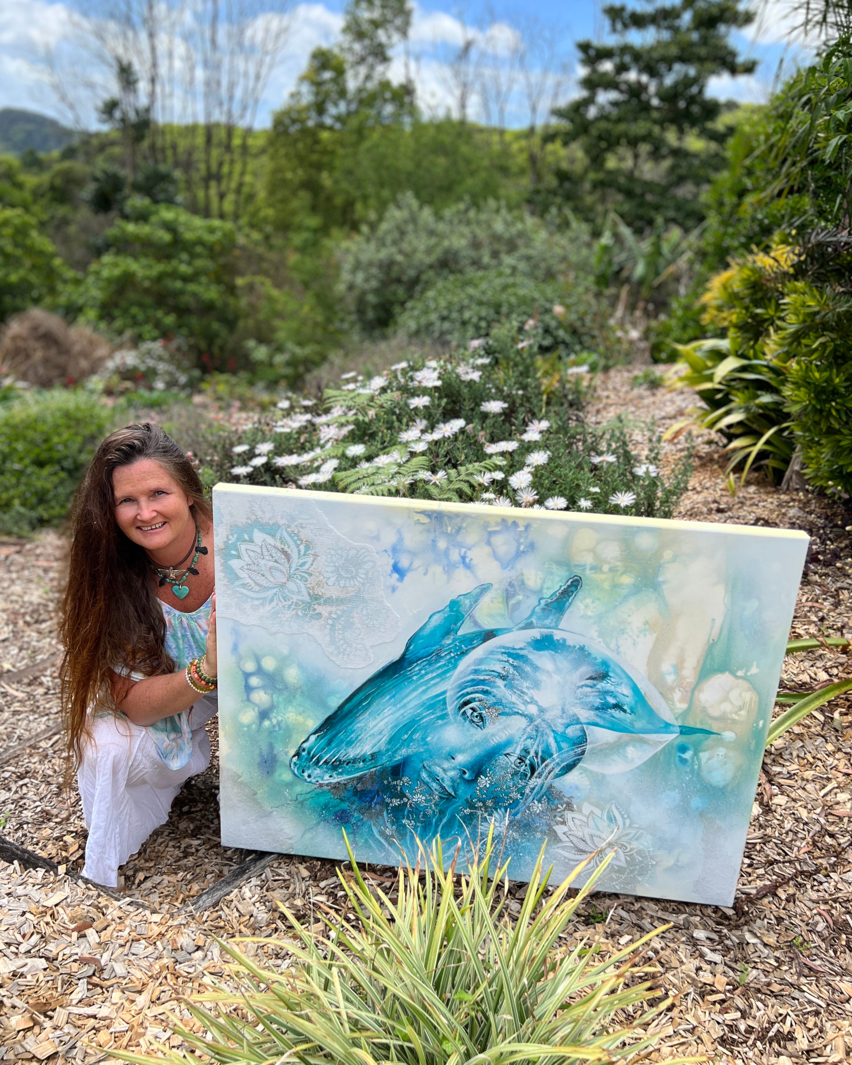 Woman holding a painting of a blue whale in a natural setting with greenery and flowers.
